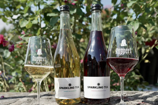 Zwei Flaschen Sparkling Tea, Grüner Tee & Rosmarin und Hibiskus & Johannisbeere, mit zwei gefüllten Weingläsern von Ruppert
