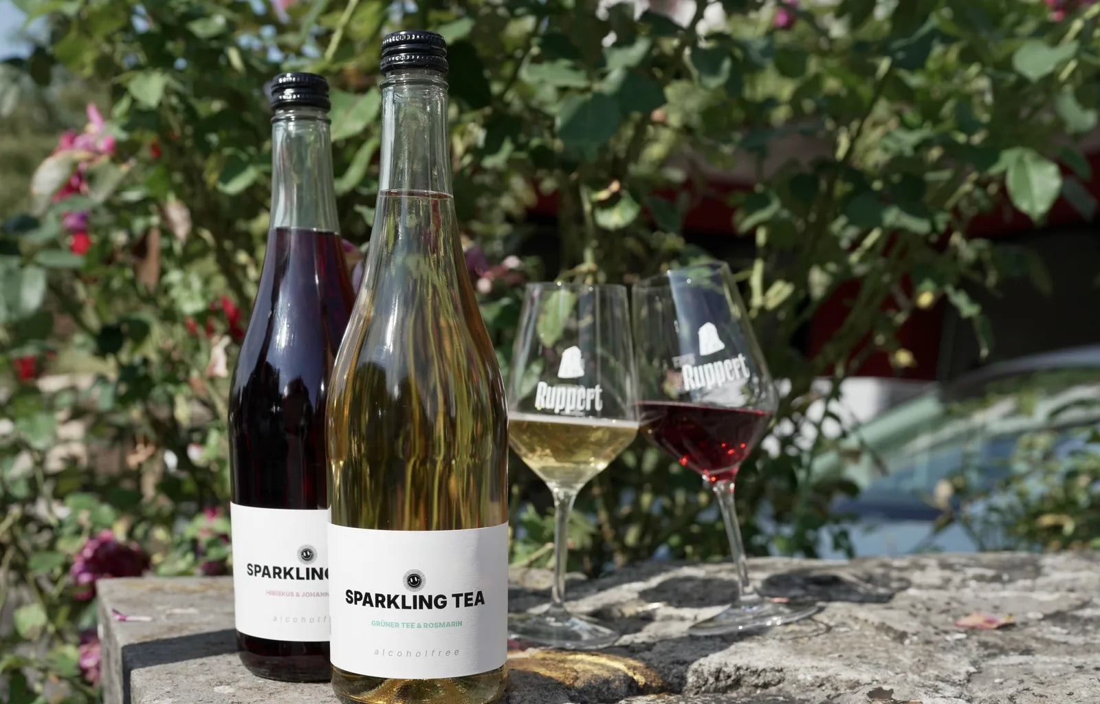 Zwei Flaschen Sparkling Tea, Hibiskus & Johannisbeere und Grüner Tee & Rosmarin, mit zwei gefüllten Gläsern vom Weingut Ruppert